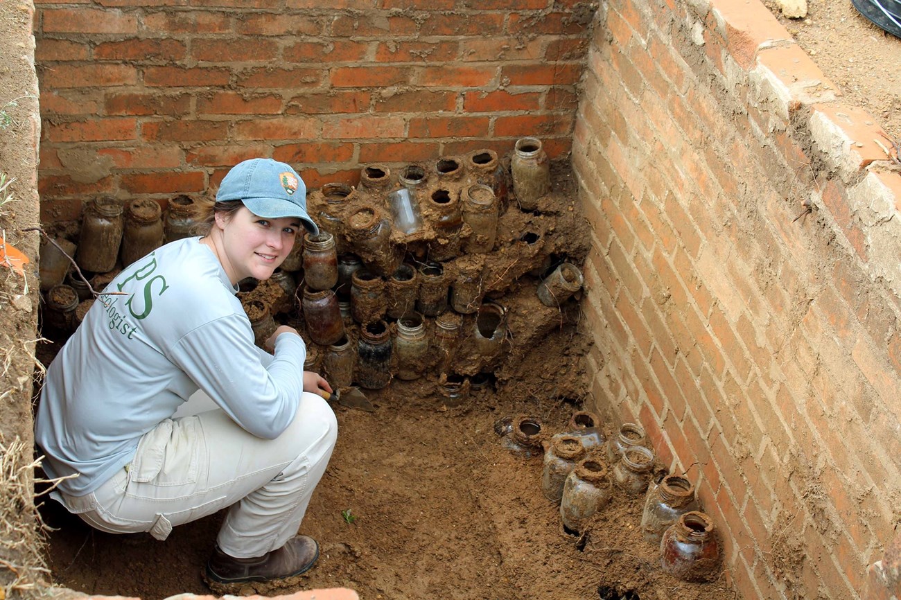 NPS archeologist kneeling next to historic glass jars at Robert E. Howard House Museum in Cross Plains, Texas NPS archeologist kneeling next to historic glass jars at Robert E. Howard House Museum in Cross Plains, Texas