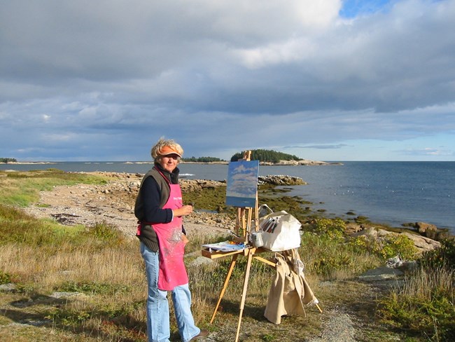 Photograph of artist Priscilla Whitlock on the coastline with her easel