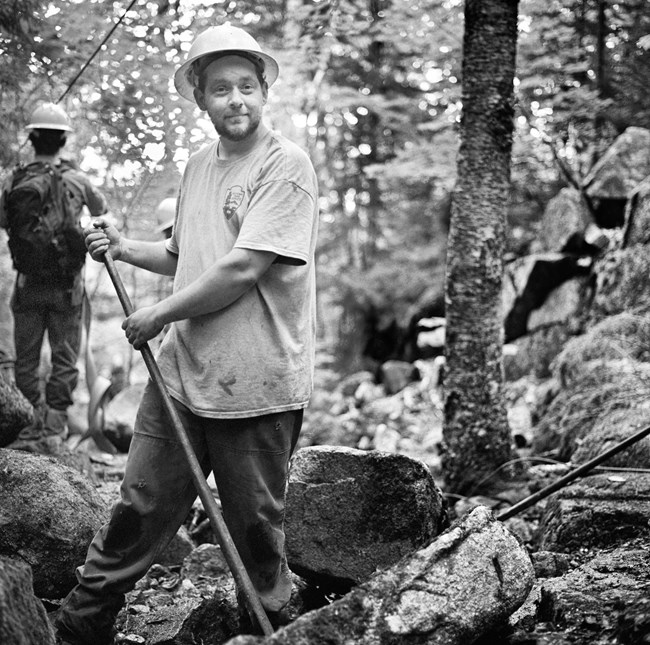 Black and White photograph of a trail crew worker using a pry bar to move large rocks.