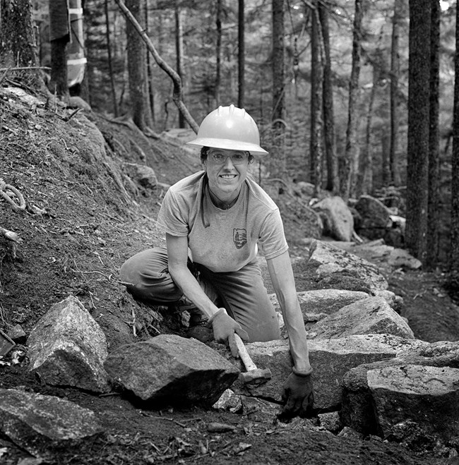 Black and White photograph of trail crew worker with a hard hat on and working on a trail. They are bent over and have a hammer in their hand.