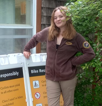 Young woman in a brown fleece with an NPS volunteer patch stands next to a row of bike safety signs