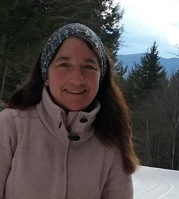 woman wears scarf and winter coat with trees and snow covered hills in background