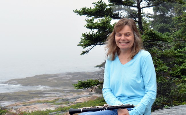 Woman with oboe on her lap smiles with trees and coastline in background