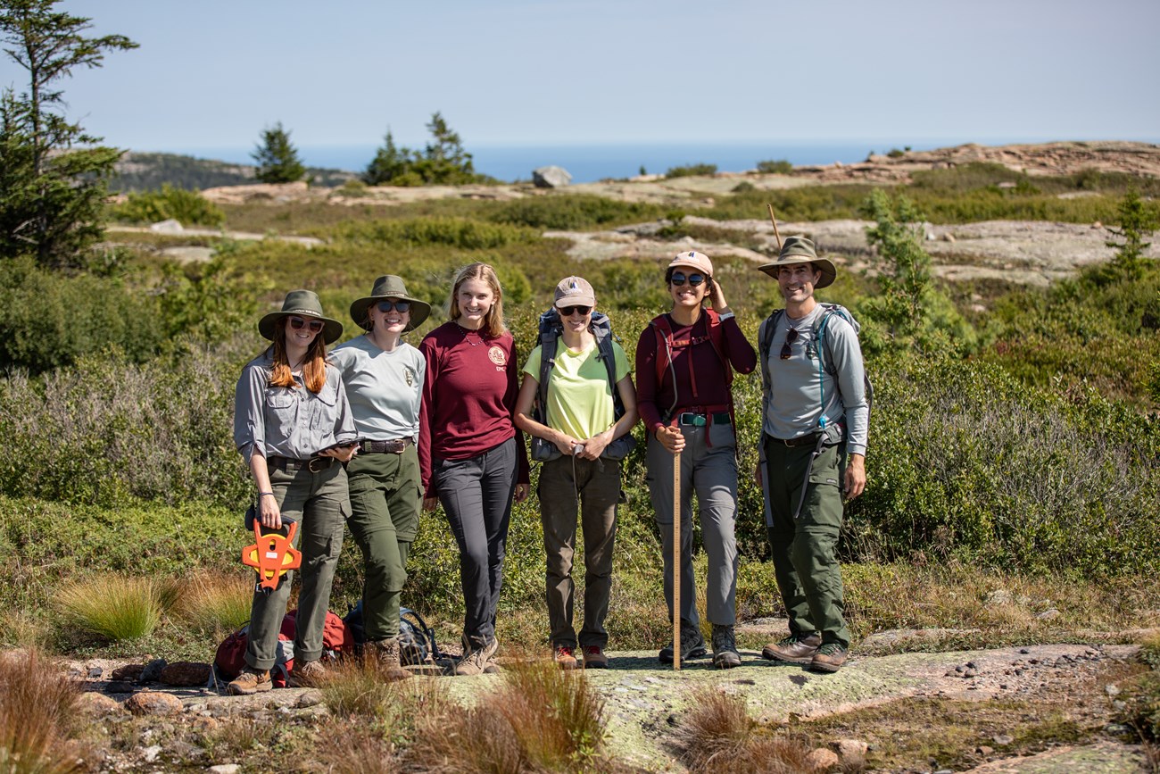 Six people in volunteer, partner, and NPS staff uniforms stand together in a summit restoration work site