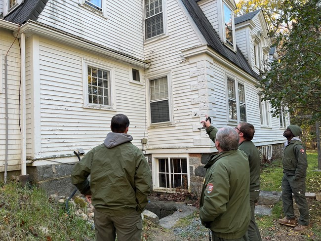 A group of uniformed NPS staff gather outside an aging white household structure for a condition assessment
