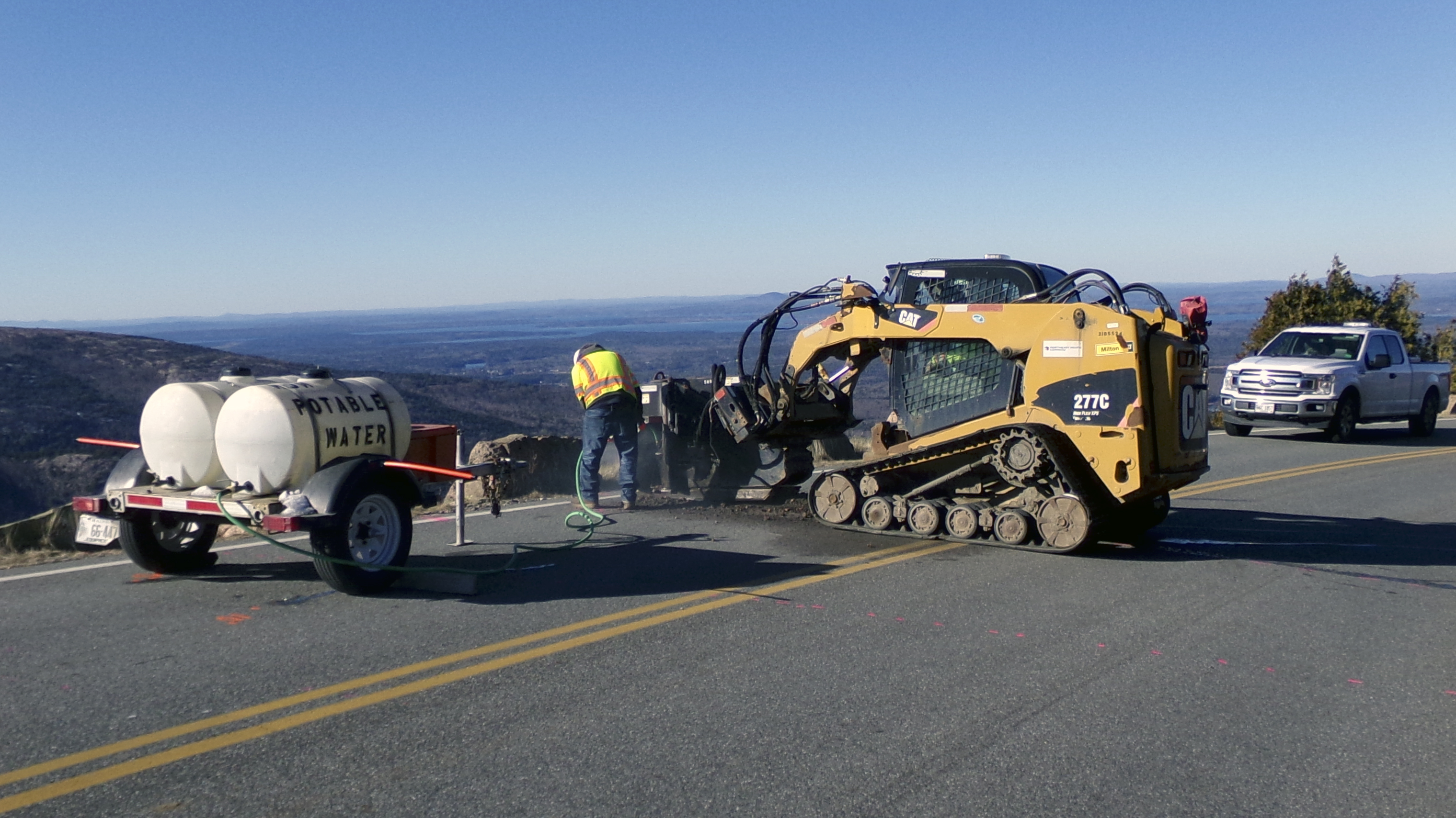 A worker in a hard hat and safety vest stands next to heavy equipment on a mountain roadway