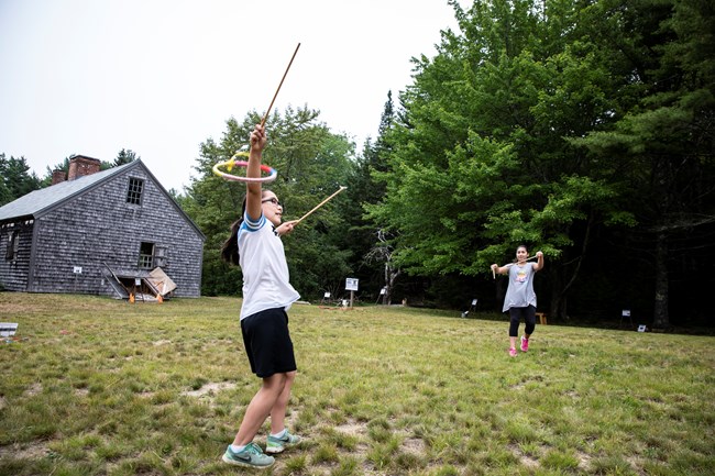 Two children are in motion holding sticks in front of a wood shingle house, while one catches a colorful hoop with one stick