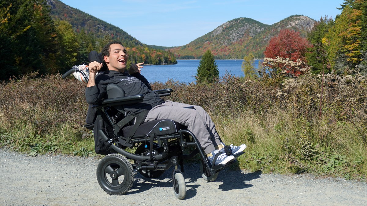 Smiling man in wheelchair parked on a path with mountains and pond behind him.