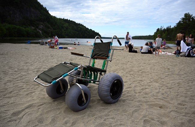 Wheelchair with large inflatable tires on a sandy beach with a lake and forested hills in the background