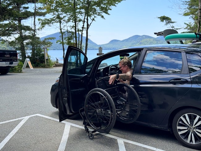 In a parking lot with mountains in the distance, a woman in a wheelchair transitions to the driver seat of a minivan