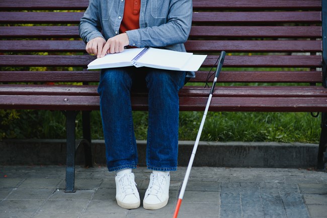 Photo of the lap of a person using a braille document while sitting on a park bench with a red-tipped can beside them