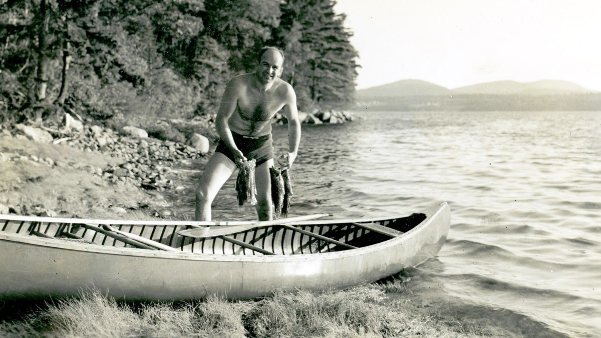 Shirtless man in swim trunks holds two batches of fish by canoe resting on the shoreline of a lake