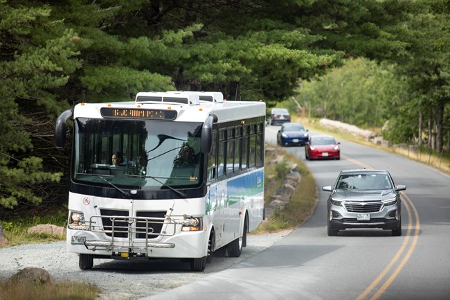 A white bus with green and white stripes is parked along a bend in a road with several cars in traffic passing beside it