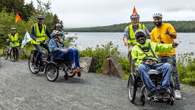 Two groups of people on special wheelchair bikes on a carriage road with a lake and mountains in the background