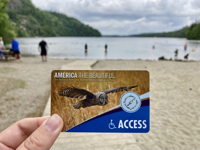 A hand holds an Access Pass card in front of a sandy lake beach