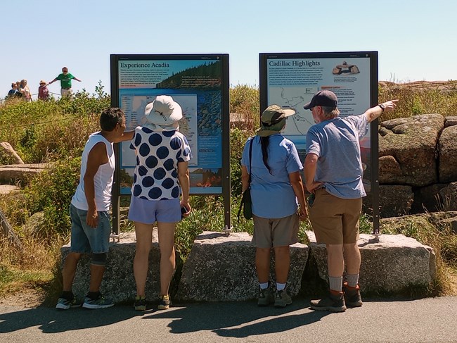 Four visitors in summer shirts, hats, and shorts read and discuss contents of two vertical orientation panels on the summit of Cadillac Mountain
