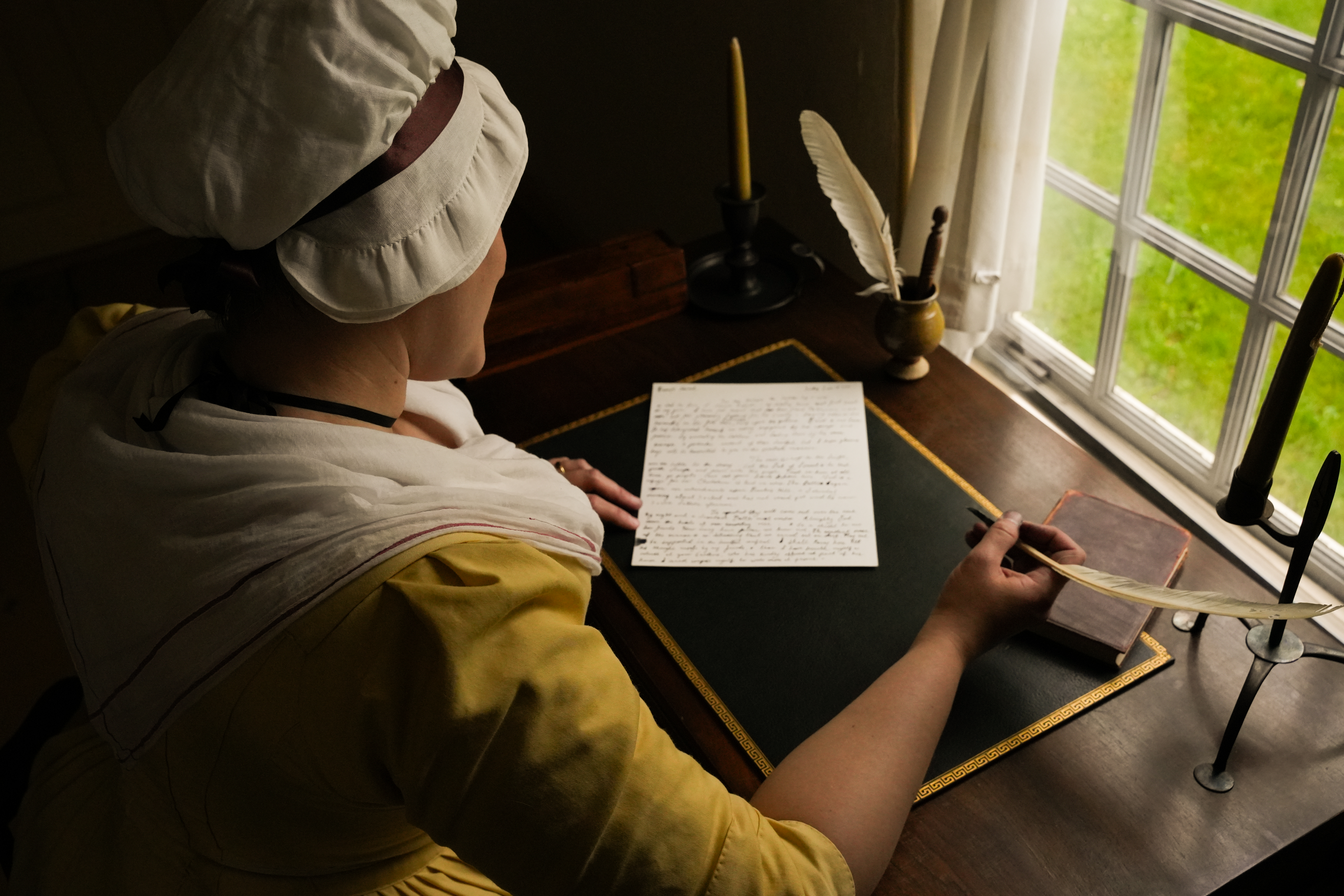 A woman wearing a dress and cap in the 1700s style sits at a desk and writes a letter with a quill pen