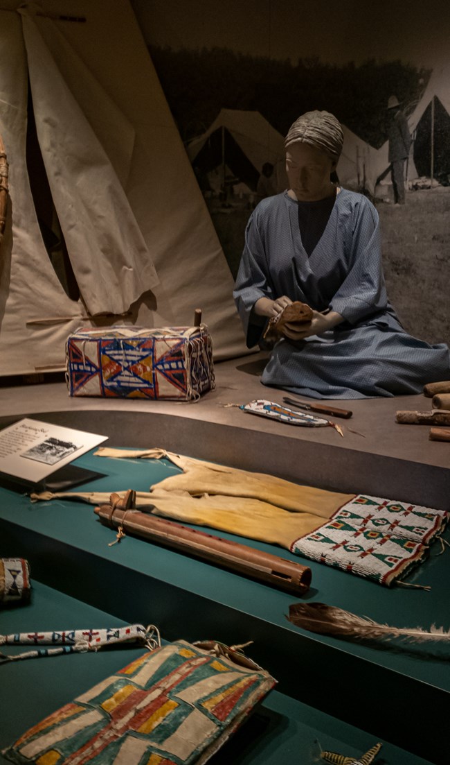 Mannequin of woman in blue dress working next to a tipi. Native American craft artifacts displayed in front of her.