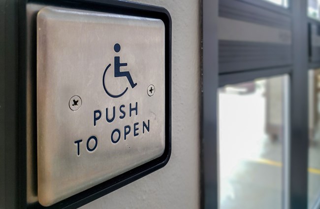 Square metal button with wheelchair logo to the left of a set of glass doors. Button reads “Push to Open”.
