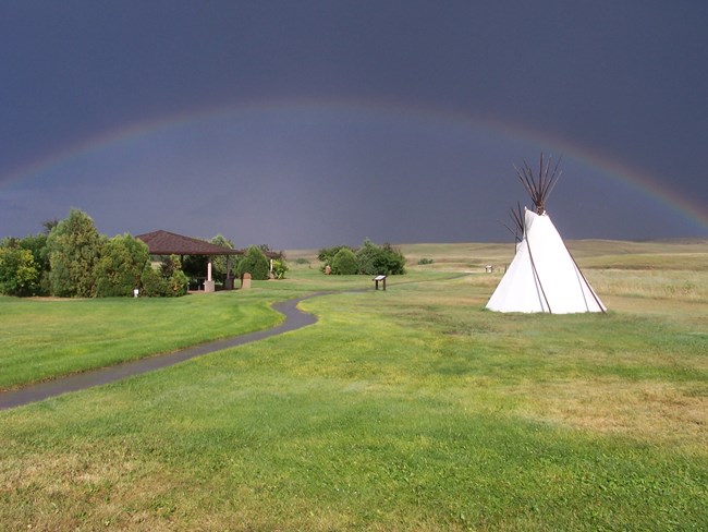 A rainbow arcs over prairie, a walkway, pavilion, and tipi.