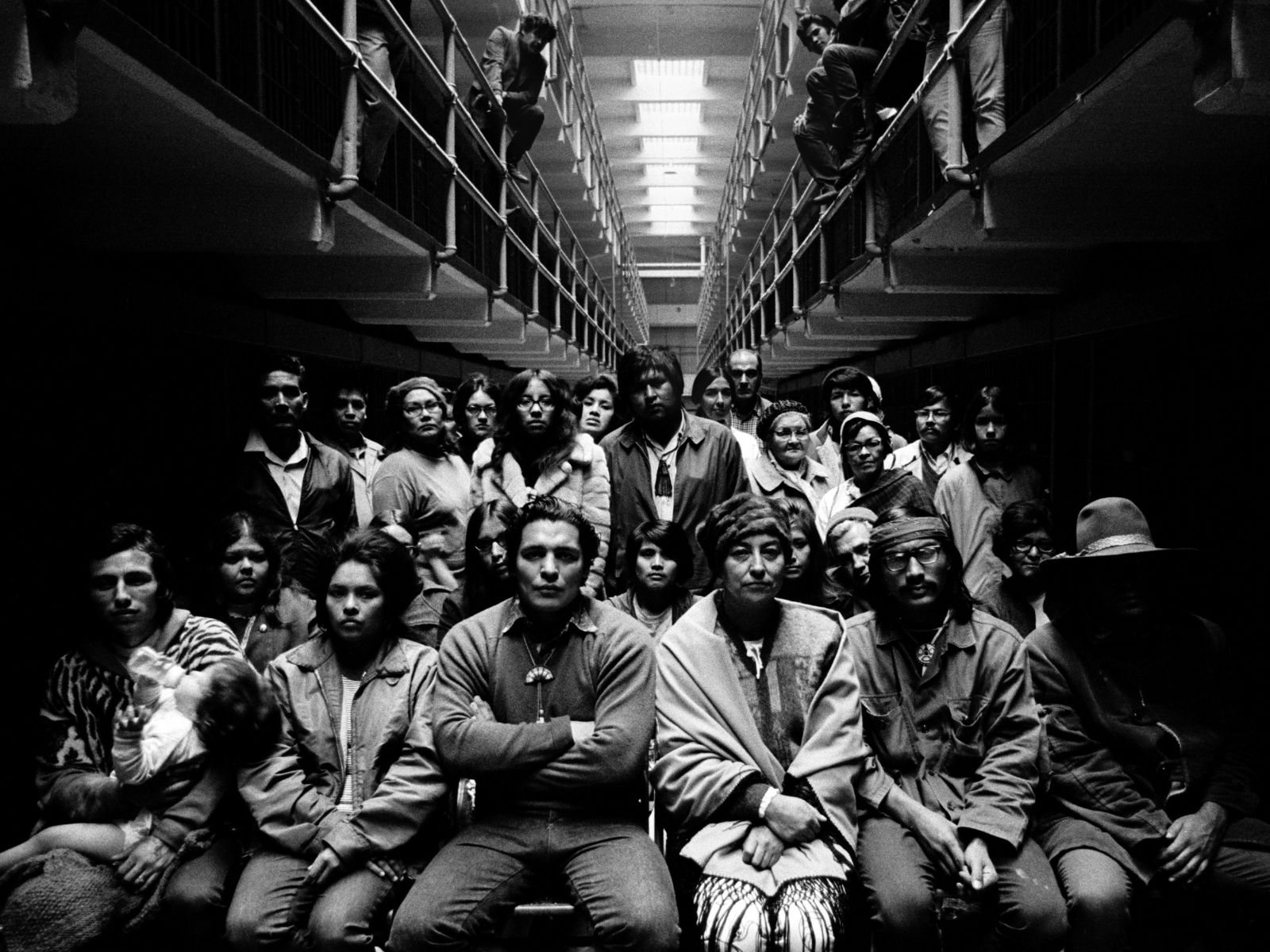 Black and white photo of large group of indigenous people, seated and standing, in the central isle of a prison block. The second tier of prison cells recede into the distance, with more people leaning on its railing, above the crowd.