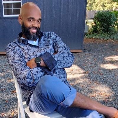Bearded African American man in blue button-down shirt sits and smiles at camera, ankle crossed over knee and arms crossed.