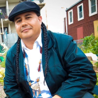 Indigenous man with braids and newsboy hat smiles at camera