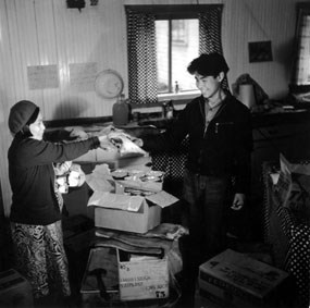 Black and white image of a woman and man standing in room cluttered with open cardboard boxes and papers.