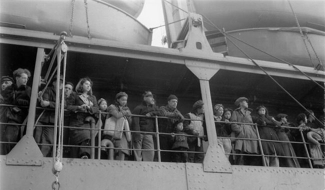 a historic scene of passengers on a boat lining the railing and looking out away from the ship.