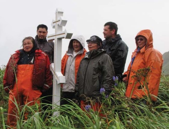 A group of people wearing rain jackets and rain pants stand around a white cross in a bright green field.