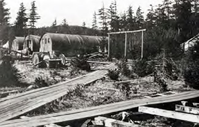 a historic scene of five long, rounded metal structures surrounded by a disarray of lumber and pathways.