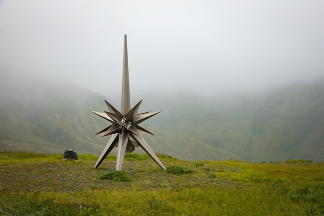 a large silver sculpture of a star with multiple points sits on a grassy slope shrouded in thick fog.