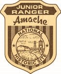 wooden junior ranger badge reads "Junior Ranger" across the top. "Amache" is in script underneath. "National Historic Site" is in a circle around a scene depicting a barrack and guard tower.