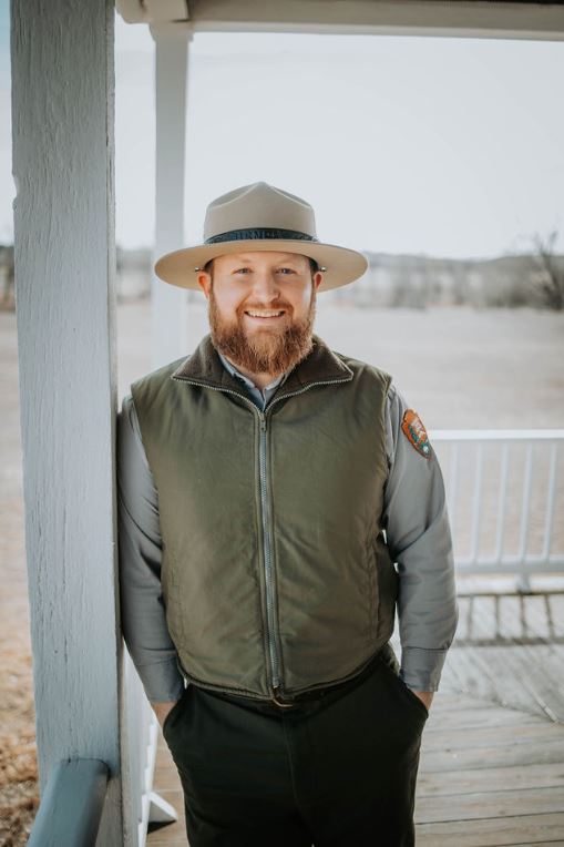Site Manager Chris Mather appears under the porch awning on a grey day with a green vest and ranger flat hat.