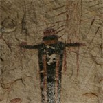 Close up of a red humanoid with arms outstretched and appearing to wear long, draping garments painted on a cave wall in front of red line paintings of other humanoids.