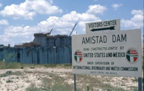 Color image of cement construction behind a sign stating "Amistad Dam being constructed jointly by the United States and Mexico under the supervision of International Boundary and Water Commission."