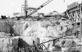 Black and white image of old crane structures near the edge of a rock cliff with high concrete forms and partial dam structure