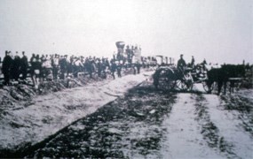 Hundreds of people lining both sides of the tracks in front of a steam locomotive next to a two-track road with a horse and wagon and men on the wagon parked across it.