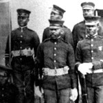 Five black soldiers at attention in dress uniform for the Union Army. One in center of back row of three is looking up and to the left while the others look straight on.