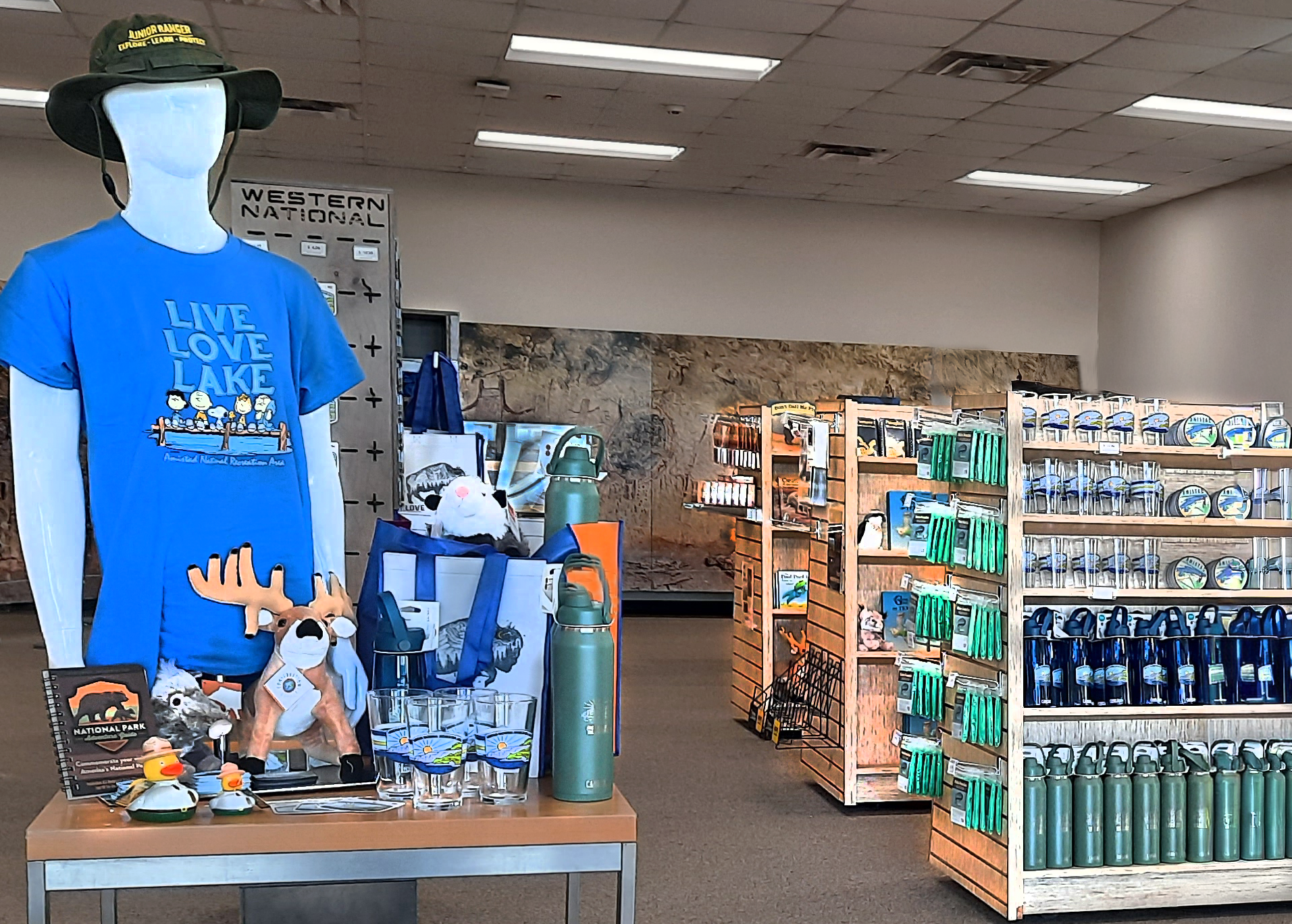 Mannikin torso wearing an Amistad National Recreation Area t-shirt and surrounded by a variety of souvenirs, and three shelf units full of books, water bottles, beverage glasses, ponchos, and other items for sale.
