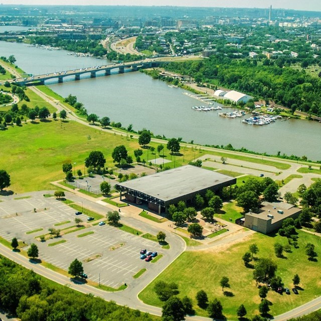 A large building with parking lots surrounding it sits along a river, with a large city in the background.