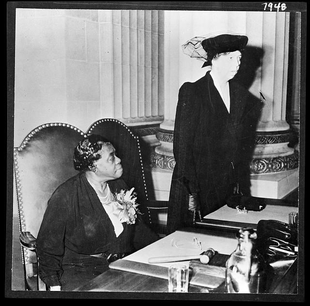 A black and white photograph of a woman is standing next to another woman seated in a chair.