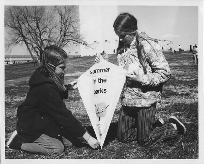 Two girls hold a kite that has "summer in the Parks" written on it. The Washington monument is in the background.