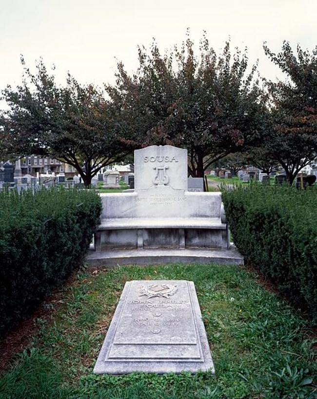 A concrete tombstone is surrounded by bushes and other graves.