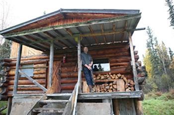 Man standing on porch of log cabin in forest.