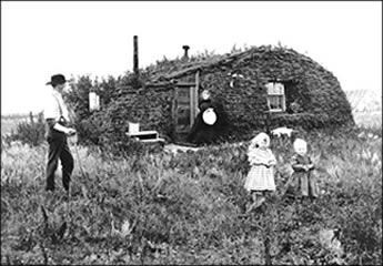Black and white photo of old sod home in open prairie with two adults and two small children.