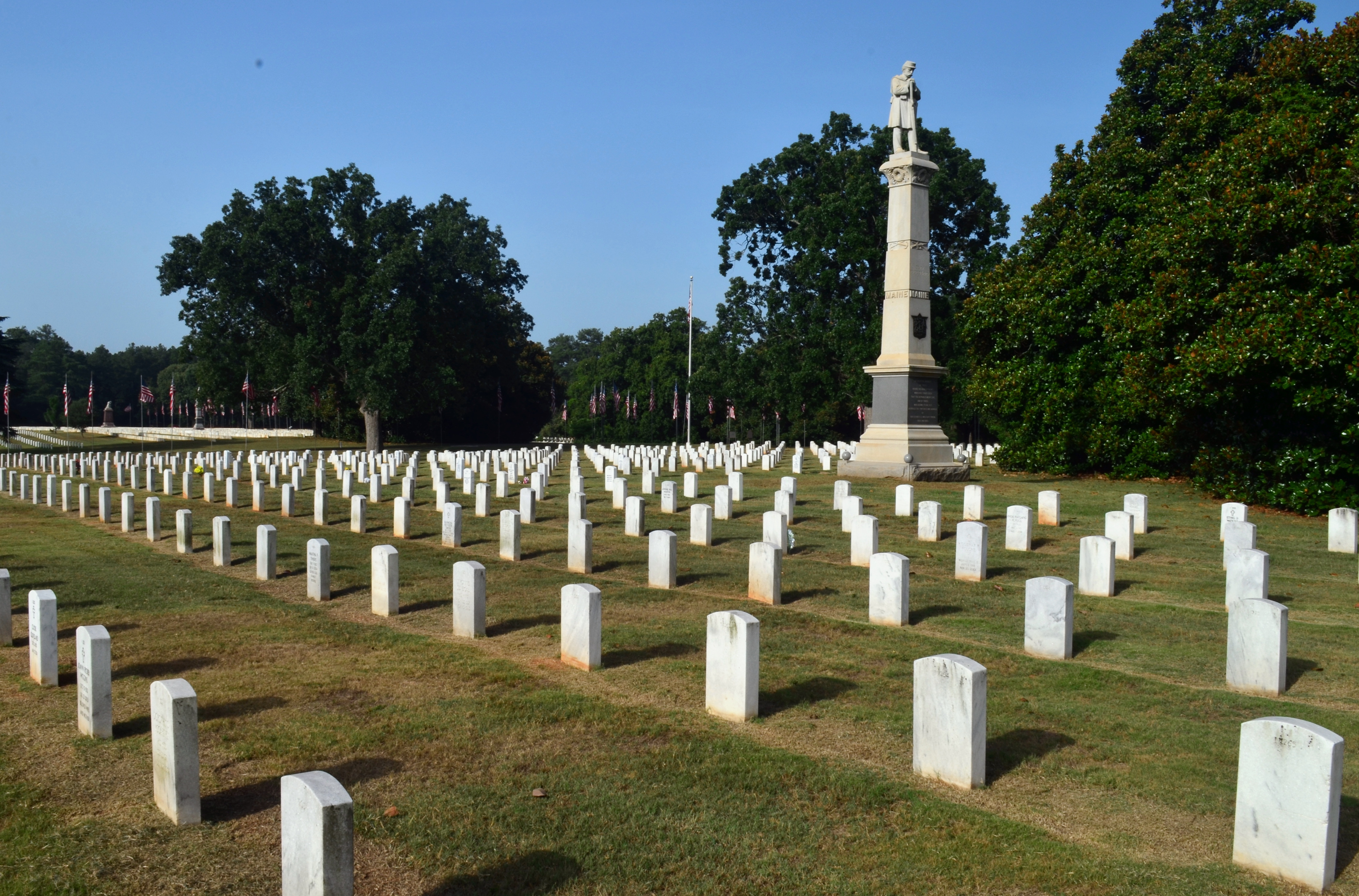 Monument in cemetery with trees and flags in background.