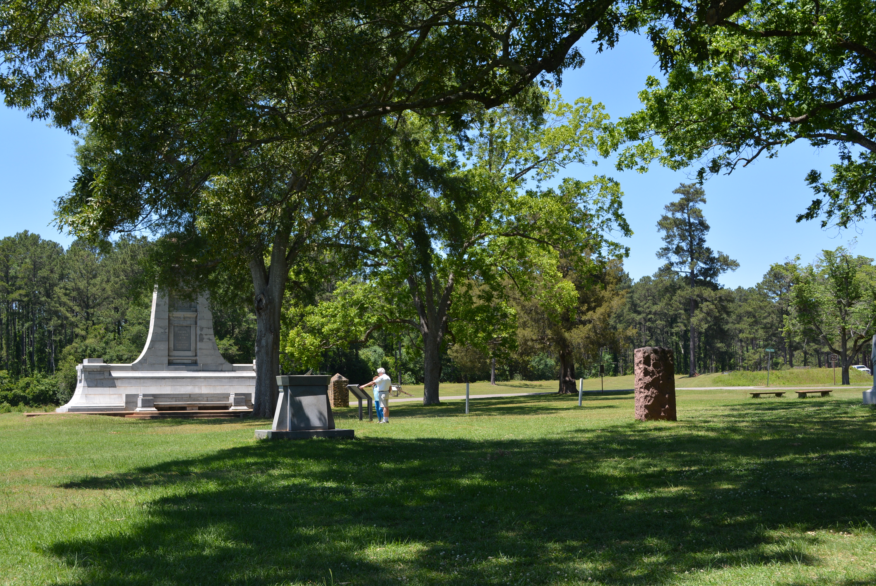 A man and woman stand near a sign in a grassy field with large trees and several large monuments