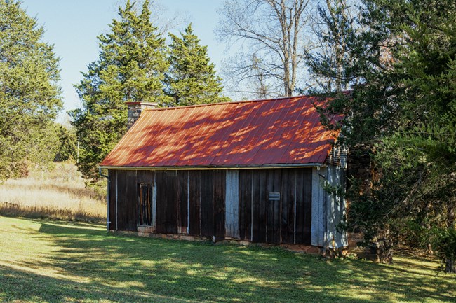 An 1790s wooden building with a red metal roof and stone chimney