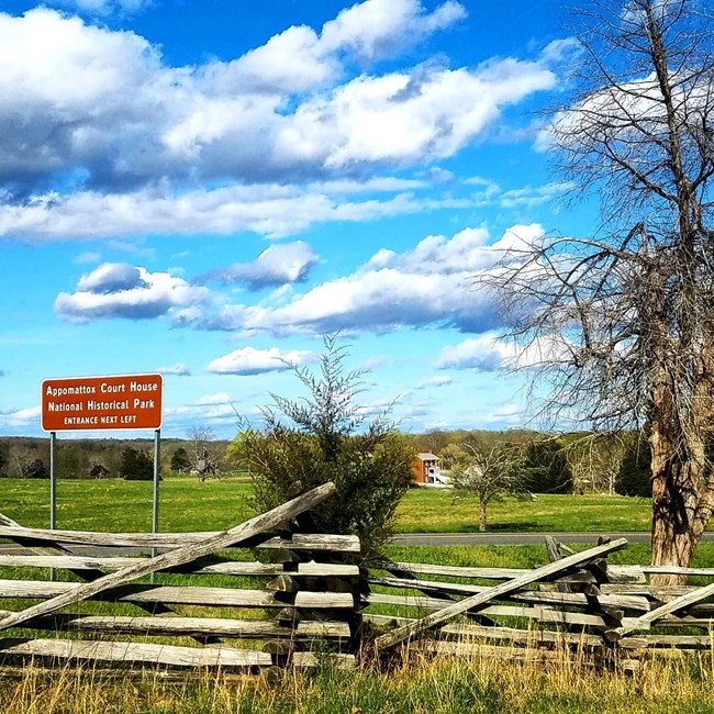A split-rail fence in the foreground with the brown directional sign for Appomattox Court House above and to the left against a deep blue sky with fluffy white clouds. The reconstructed brick McLean House is just visible on the far ridgeline.